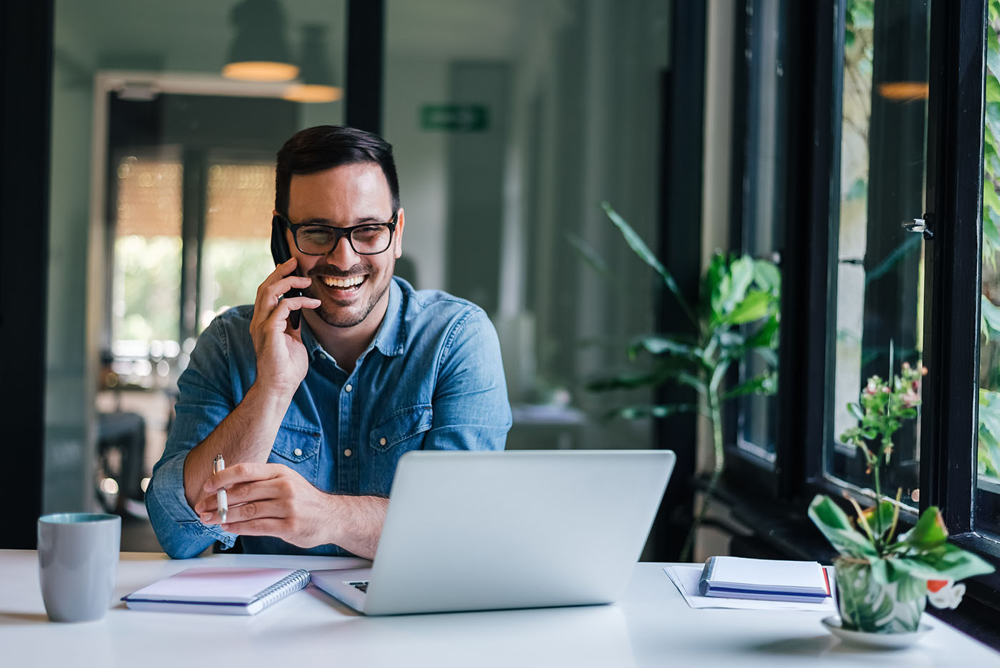 Portrait of young business man smiling in a casual office making phone call while working with laptop