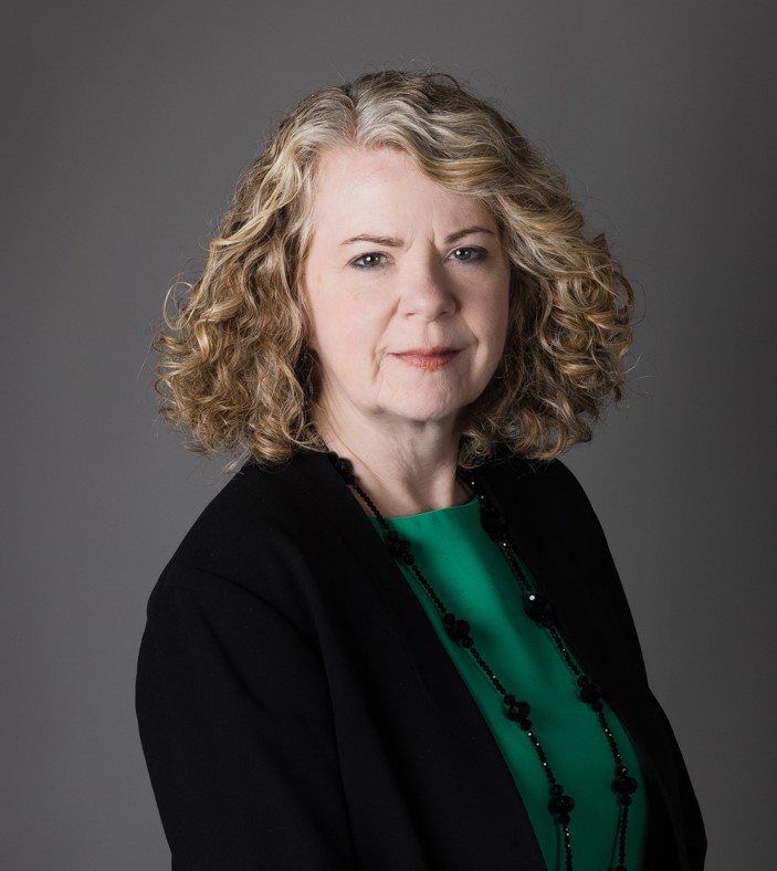 This image of a woman in a blazer with a green shirt. She has curly hair to the shoulders that has highlights around the part on the right side of her hair. She has a gaze direct at the camera and a soft smile.